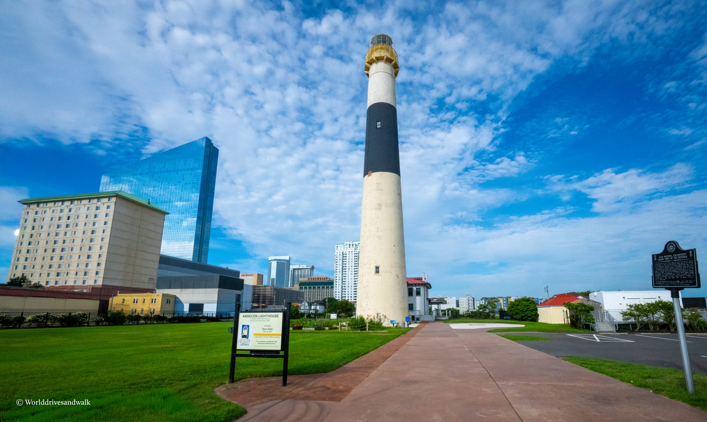A tall striped lighthouse with a golden lantern room stands on a brick pathway in a green park, with modern Atlantic City buildings visible behind it under a blue sky with white clouds.