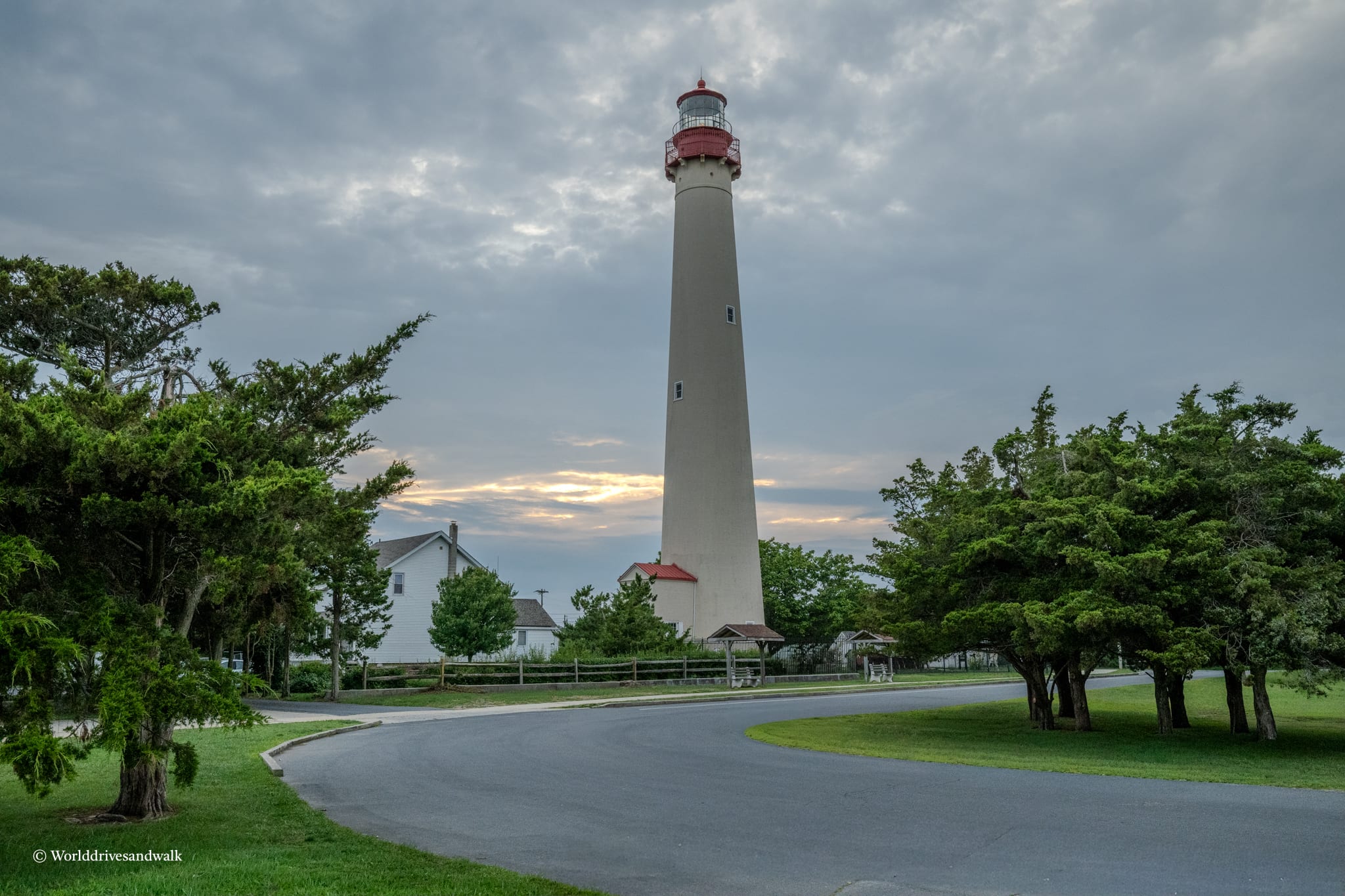 A tall white lighthouse with a red-topped lantern room stands in a grassy park surrounded by mature trees. A keeper's house with a red roof is visible to the left. The sky is overcast with breaks of golden light near the