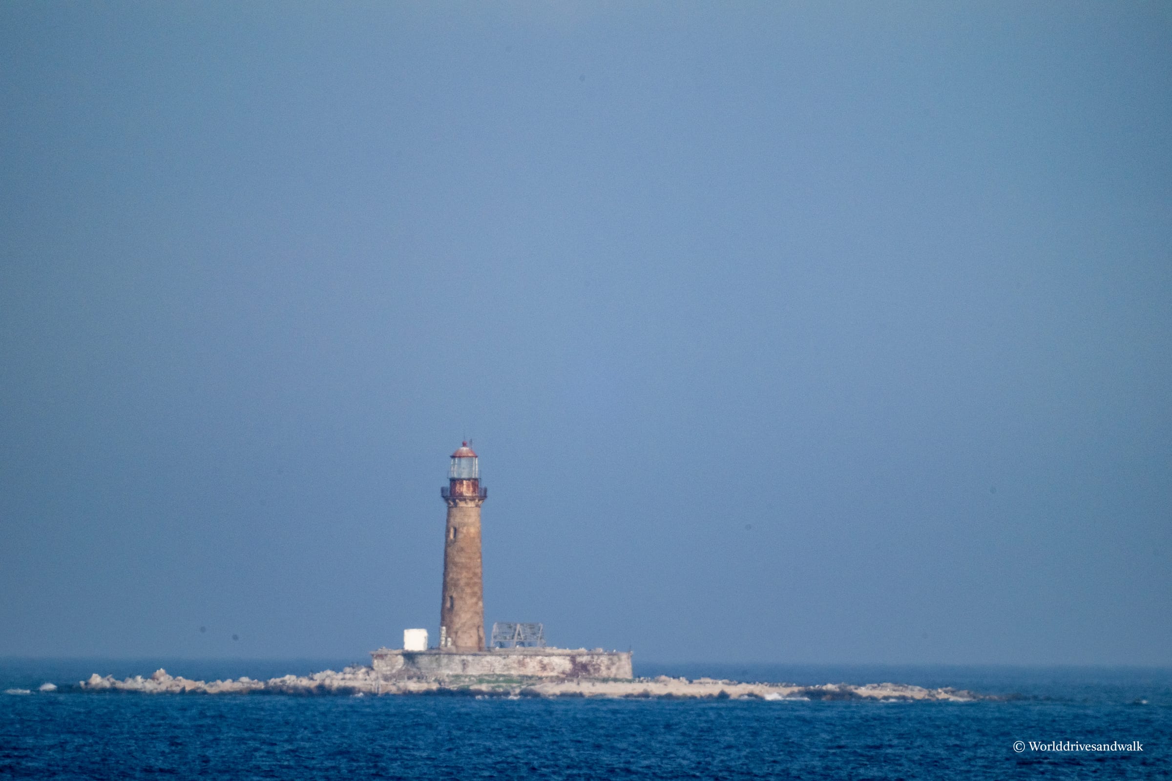 A tall stone lighthouse with a red lantern room stands on a rocky foundation surrounded by water under a clear blue sky.
