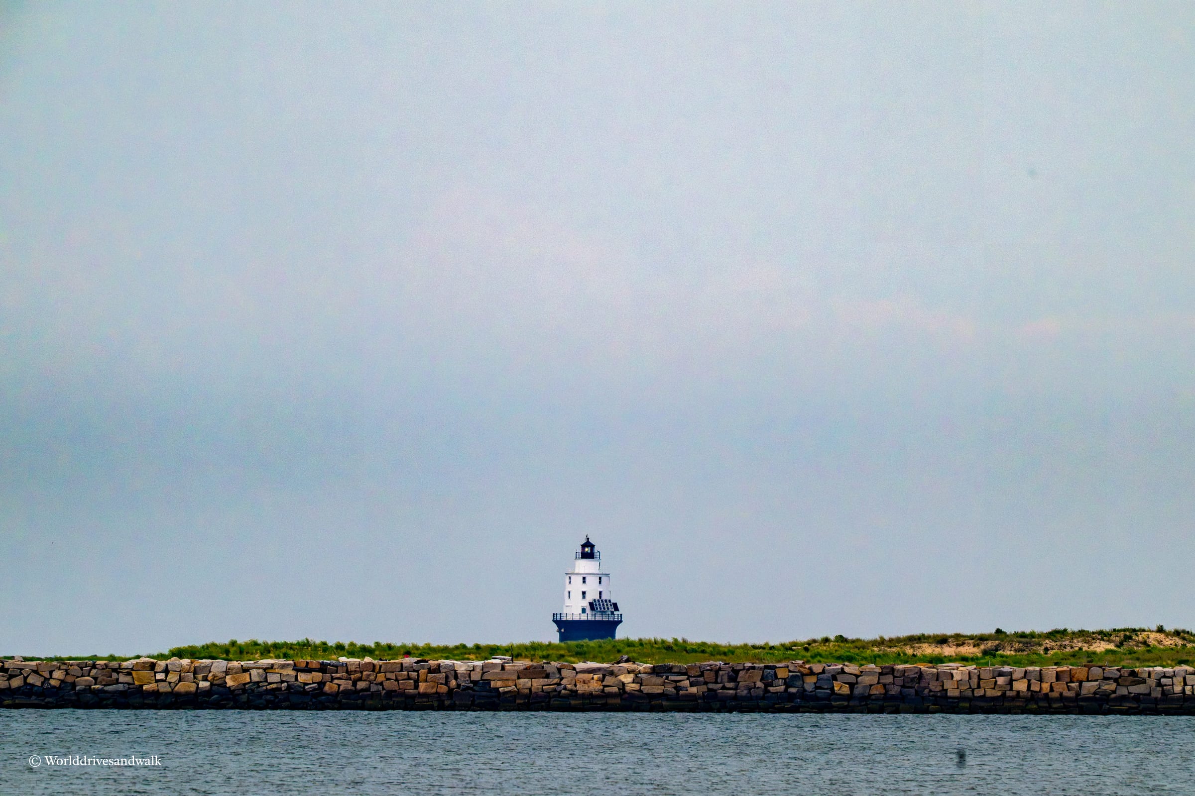 A white lighthouse with a dark lantern room stands on a grassy island behind a long stone breakwater. The structure is viewed from across calm blue-green water under an overcast sky.