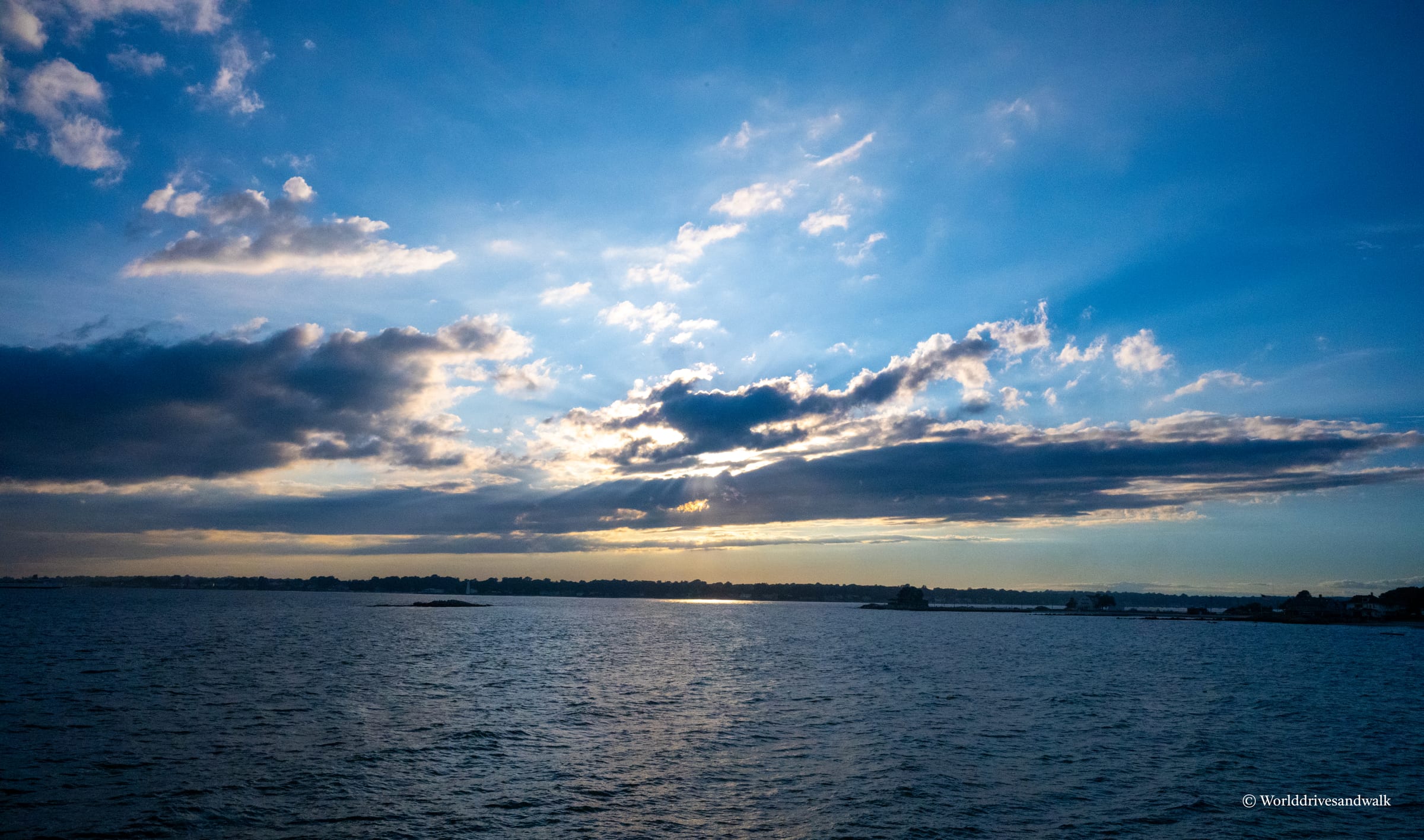 A wide expanse of calm water under a blue sky with white and gray clouds, tree-lined shoreline visible in the distance, late afternoon light filtering through cloud formations.
