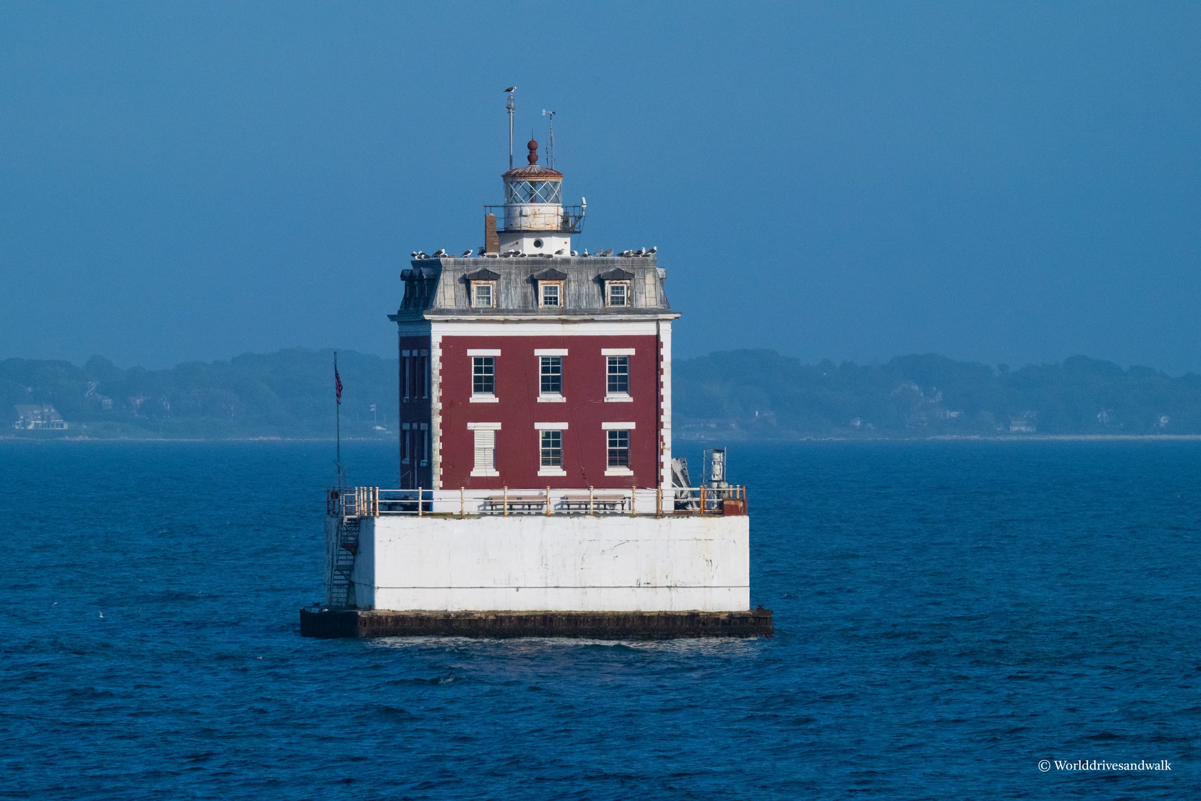 A red brick lighthouse with white trim and a mansard roof topped by a lantern room stands alone in blue water, with distant Connecticut shoreline visible across the sound.