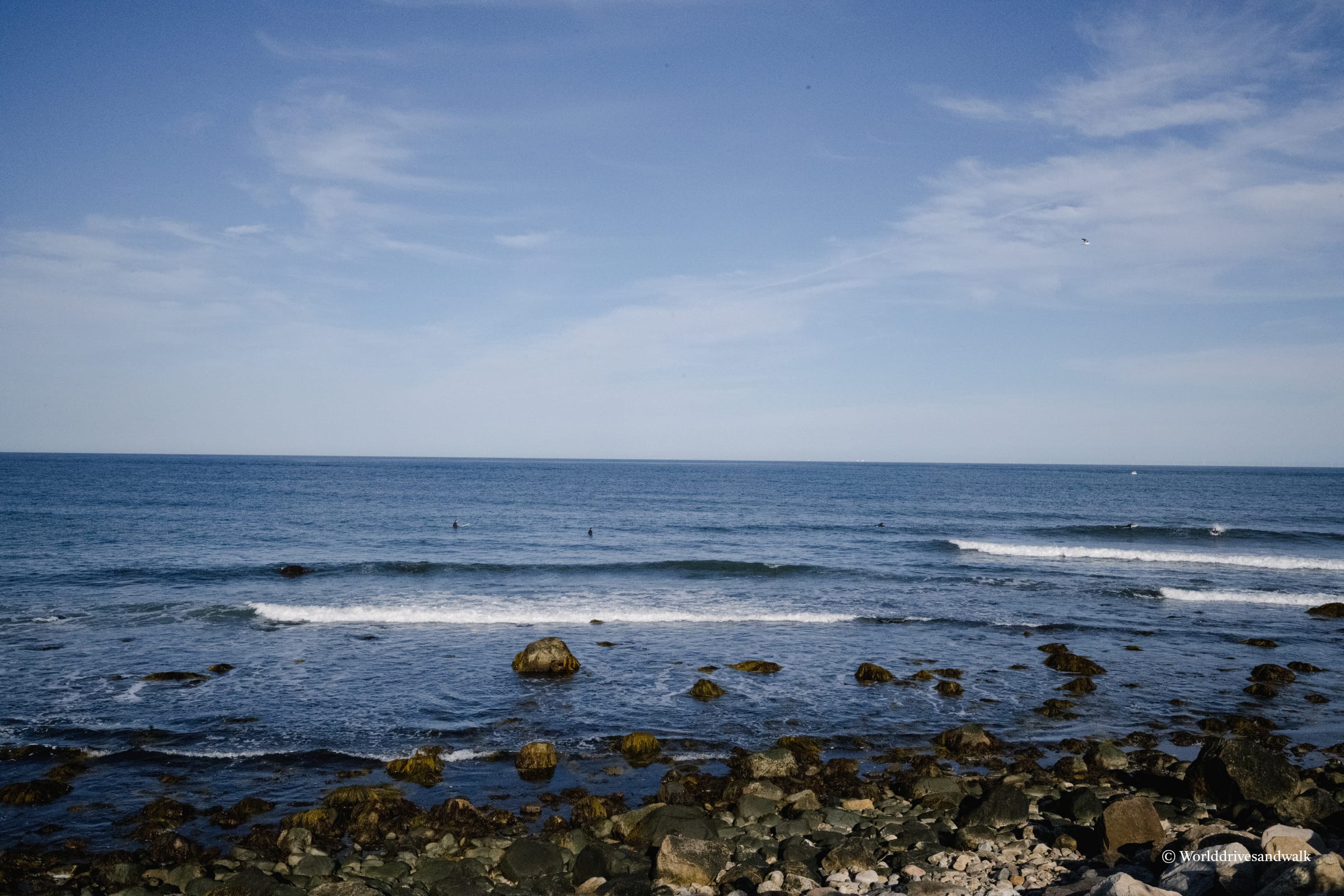Rocky shoreline with scattered boulders in foreground, calm blue ocean with gentle waves, a few swimmers visible in the distance, clear sky with light clouds.