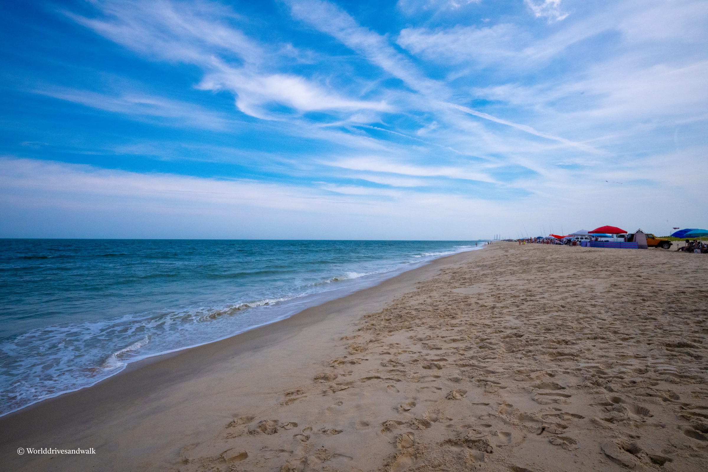 A sandy beach with footprints, a blue ocean, and a clear blue sky with scattered clouds. Several colorful umbrellas are set up on the beach.