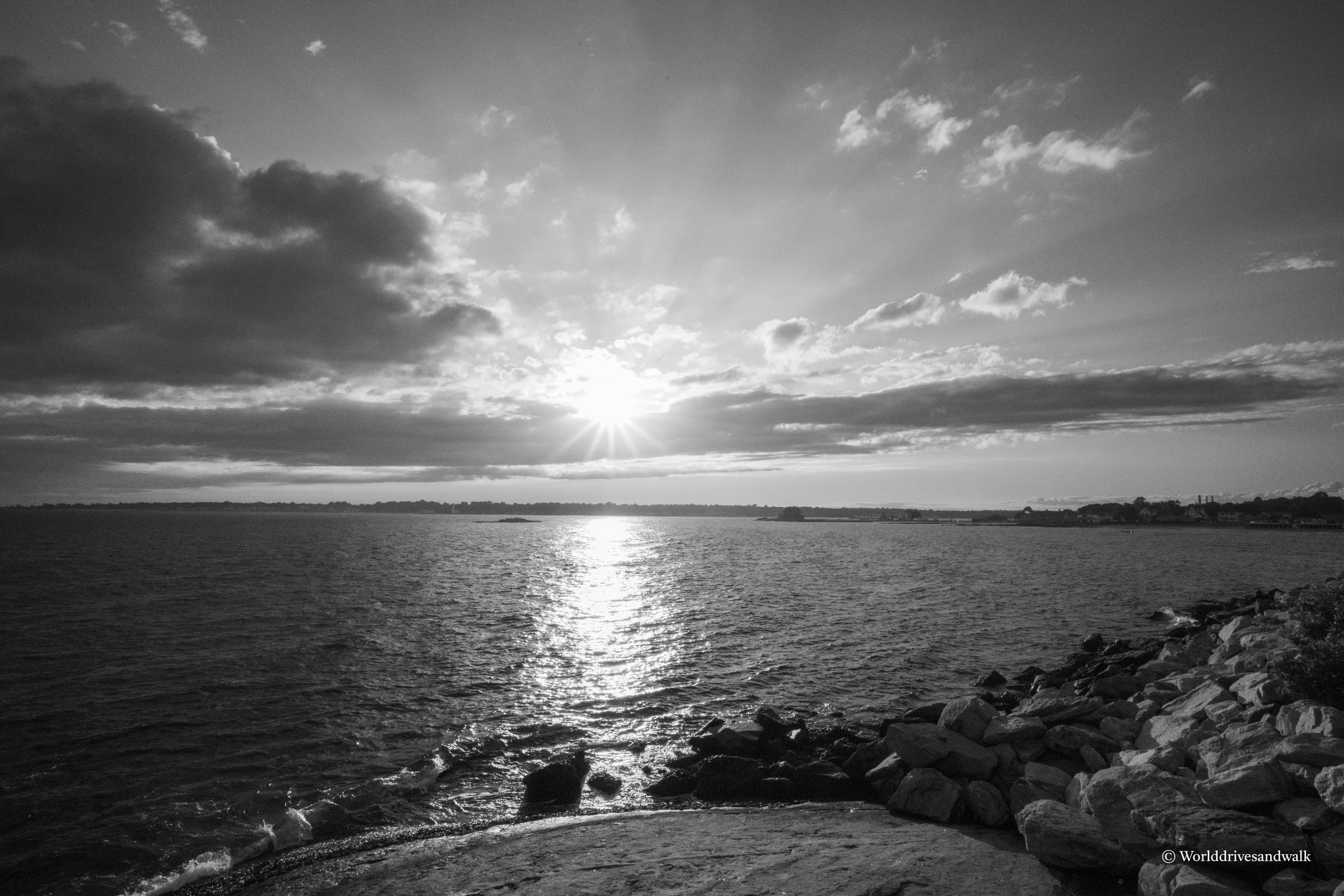 Black and white photograph of a sunset over Long Island Sound with dramatic cloud formations, rocky shoreline in foreground, and sun breaking through clouds on the horizon.